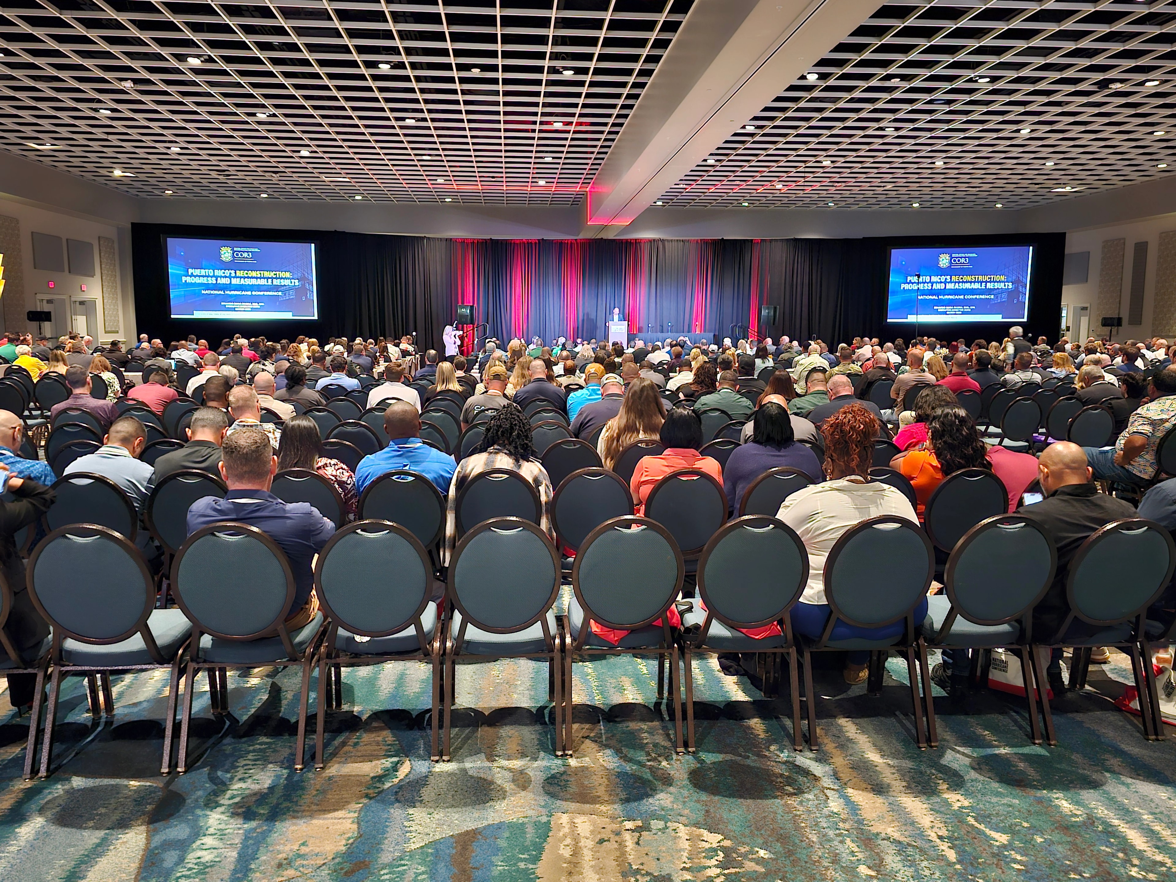 Vista panorámica del salón principal del National Hurricane Conference en Orlando, Florida, con cientos de asistentes sentados frente a un escenario donde un presentador habla ante dos pantallas grandes que muestran la presentación de COR3 sobre la reconstrucción de Puerto Rico.