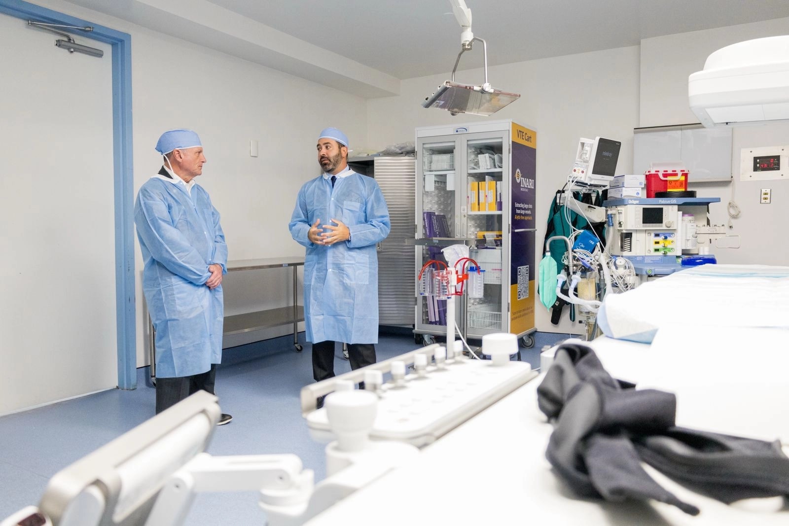 Dos hombres vestidos con batas quirúrgicas azules y gorros de sala de operaciones conversan en una sala de procedimientos del Professional Hospital Guaynabo. Al fondo se observa un carro VTE de Inari Medical y equipo médico especializado.